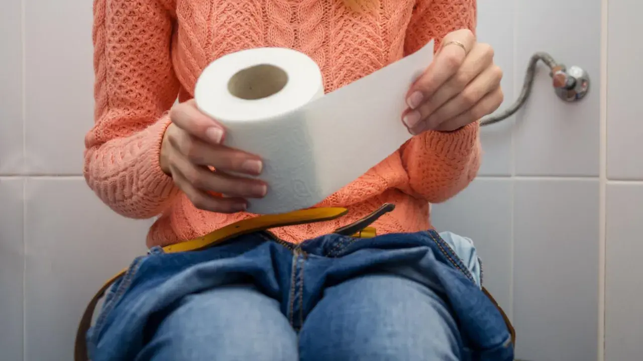 Kobieta w pomarańczowym swetrze siedzi na toalecie, trzymając rolkę papieru toaletowego. Dżinsy opuszczone, czuje, że nie może się wysikać do końca.