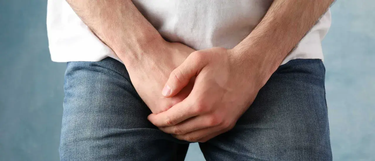 Mężczyzna w dżinsach i białej koszulce, ręce skrzyżowane na wysokości krocza. Może to symbolizować dyskomfort lub problem z popuszczaniem moczu u mężczyzn po oddaniu moczu.