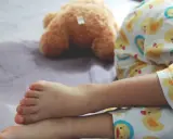 Nocne wpadki zdarzają się nawet 5-latkom. Dziecko w piżamce z kaczuszkami śpi obok pluszowego misia, a na pościeli widać mokrą plamę.