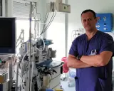 Lekarz w niebieskim uniformie stoi na tle aparatury medycznej. Jeśli masz ból jądra, to właśnie do takiego specjalisty powinieneś się zgłosić.