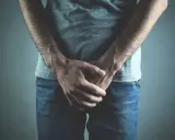 Mężczyzna w dżinsach i szarej koszulce, ręce skrzyżowane na kroczu. Może to symbolizować dyskomfort lub problemy zdrowotne, takie jak wodniak jądra.