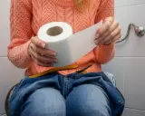 Kobieta w pomarańczowym swetrze siedzi na toalecie, trzymając rolkę papieru toaletowego. Dżinsy opuszczone, czuje, że nie może się wysikać do końca.