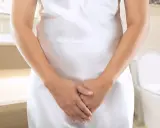 Kobieta w białej sukience w łazience, z dłońmi skrzyżowanymi na brzuchu. W tle toaleta. Szuka domowych sposobów na nietrzymanie moczu.