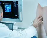 Lekarz wykonuje badanie dopplerowskie żył na nodze pacjentki, analizując obraz na monitorze.