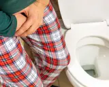 Mężczyzna w piżamie z kratę trzyma się za krocze, siedząc obok toalety. Czy zwapnienie prostaty to rak?