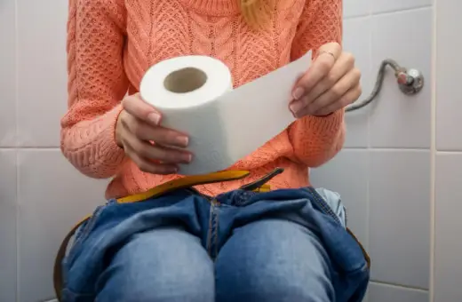 Kobieta w pomarańczowym swetrze siedzi na toalecie, trzymając rolkę papieru toaletowego. Dżinsy opuszczone, czuje, że nie może się wysikać do końca.