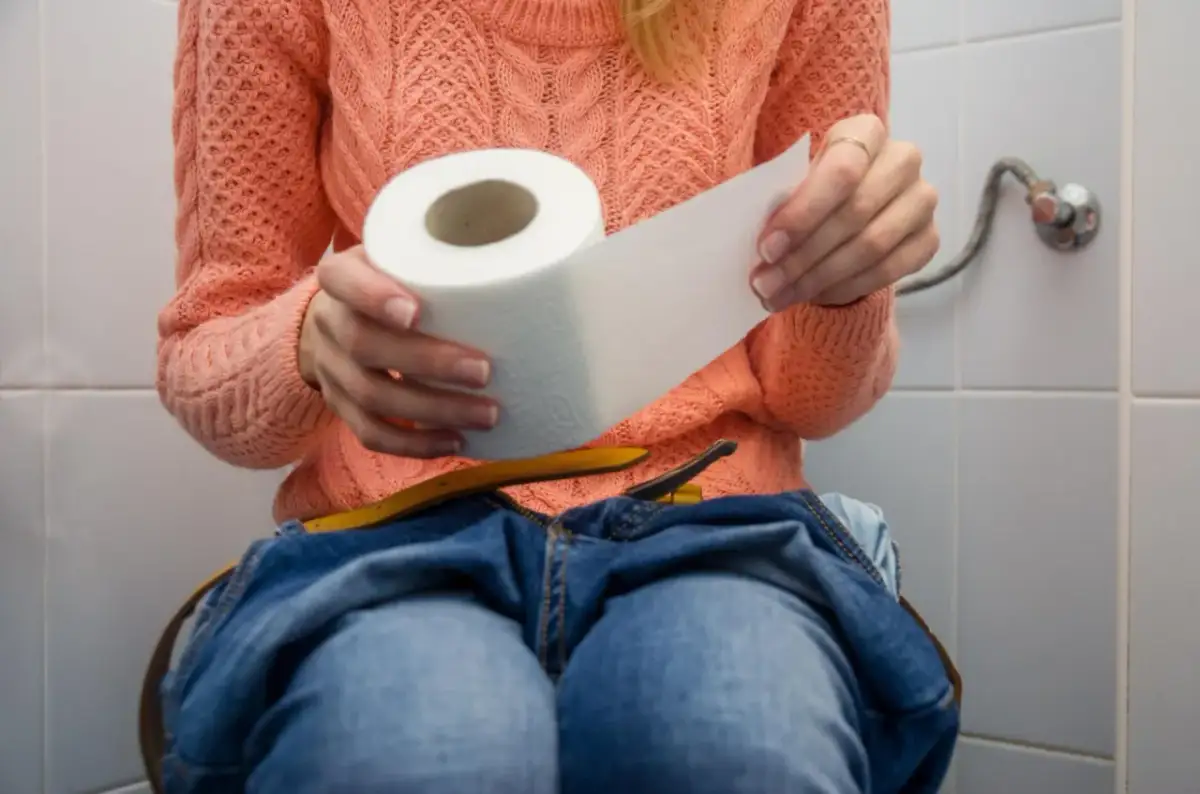Kobieta w pomarańczowym swetrze siedzi na toalecie, trzymając rolkę papieru toaletowego. Dżinsy opuszczone, czuje, że nie może się wysikać do końca.